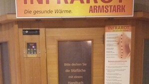 Una sauna con un letrero que dice 'INFRAROT ARMSTARK Die gesunde Wärme'. Se ven instrucciones para abrir la puerta con una llave.