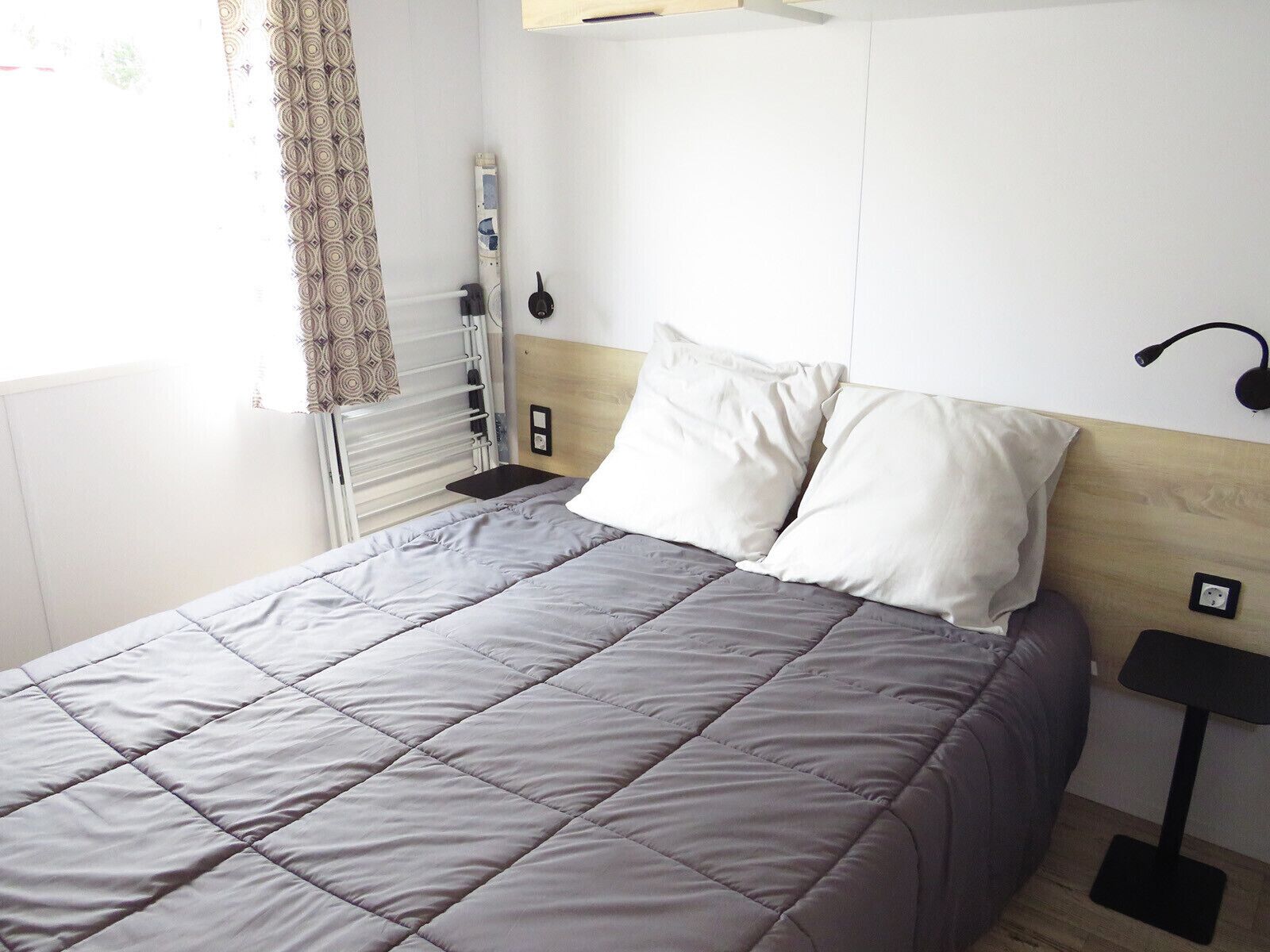 Una cama bien hecha con un edredón gris, almohadas blancas y una mesita de noche con una lámpara.