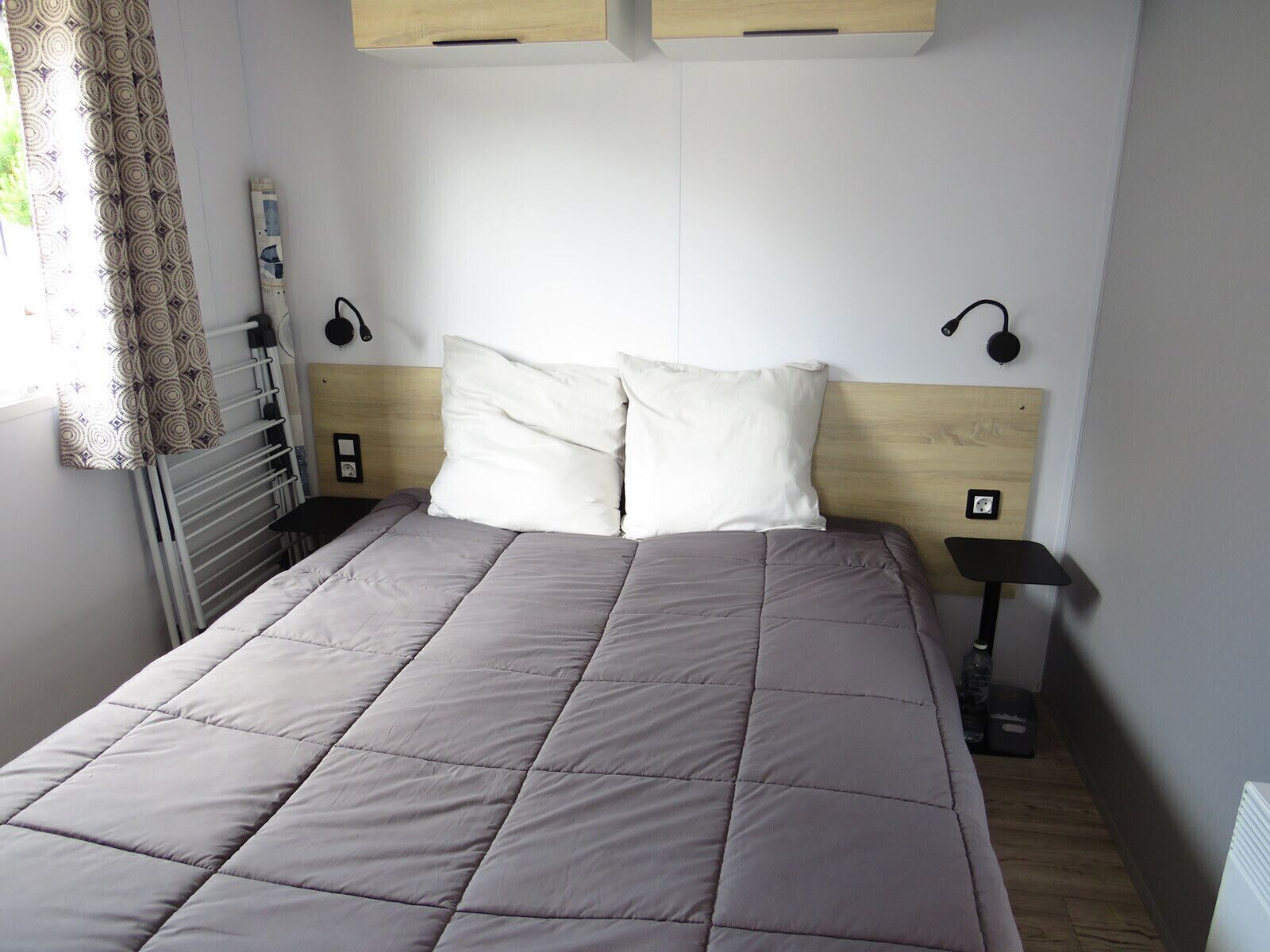 Un lit bien fait, avec une couverture grise, une tête de lit en bois et deux lampes de chevet.