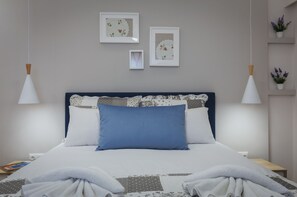 Una cama bien hecha con un almohadón azul, ropa de cama blanca con estampado, y un cabecero con diseño floral. Se observan dos marcos de cuadros en la pared, uno más grande y otro más pequeño, y una pequeña estantería con plantas en macetas a la derecha.