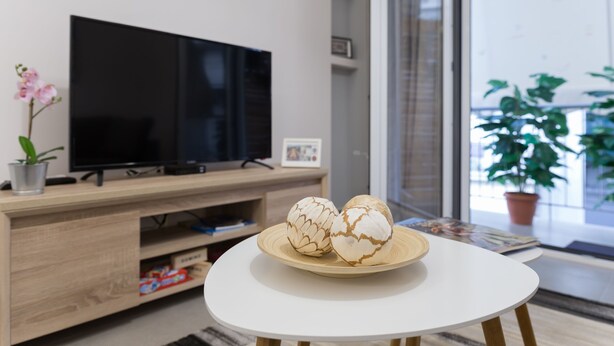 A modern living room with a flat-screen TV on a wooden stand, a round white table with decorative eggs, a potted plant, and a view through a glass door. - 57m² Koukaki Luxury Flat next to Acropolis & Metro (Athens)
