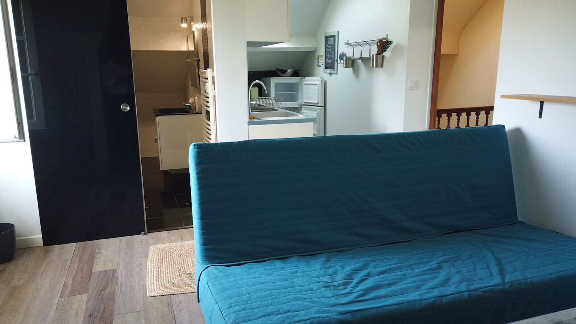 A room with a teal sofa, a small kitchenette, and a bathroom visible through an open door.