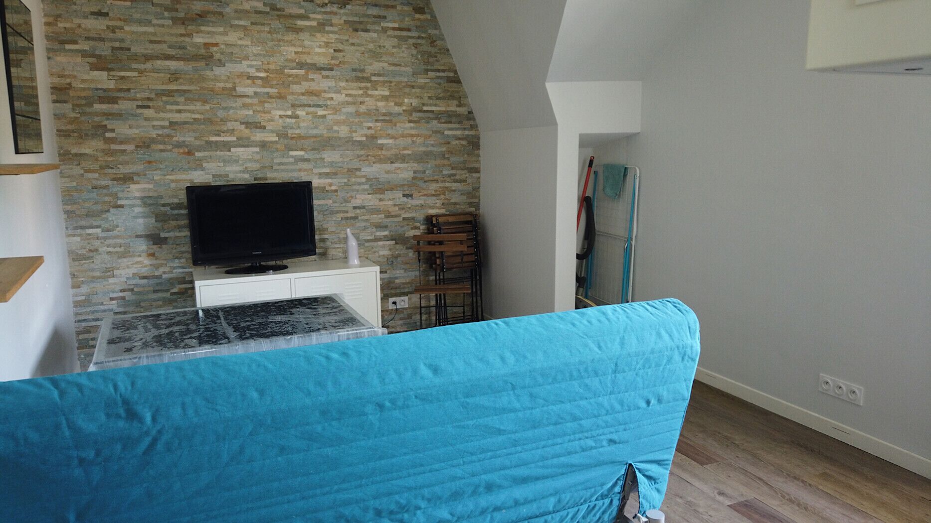 Un salon avec un mur en pierre, un téléviseur posé sur un meuble blanc, un canapé bleu et une chaise en bois.