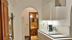 Un couloir étroit mène à une cuisine dotée d’un îlot central, d’un plancher en bois et d’une salle de bain visible au fond.