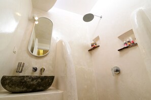 A bathroom with a stone basin sink, a mirror, a showerhead, and shelves with toiletries.