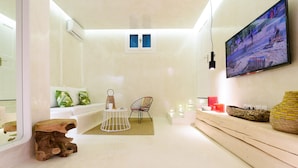 A modern, well-lit room with a sofa, a chair, a small table with a lamp, a TV mounted on the wall, and a decorative wall shelf. - Mykonos 52m² Luxury Apartment Sea side Ornos (Mykonos)