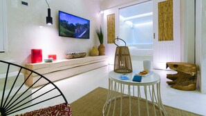 A modern living room with a flat-screen TV mounted on the wall, a round white table with a laptop and a glass, and a large window with bamboo curtains. - Mykonos 52m² Luxury Apartment Sea side Ornos (Mykonos)