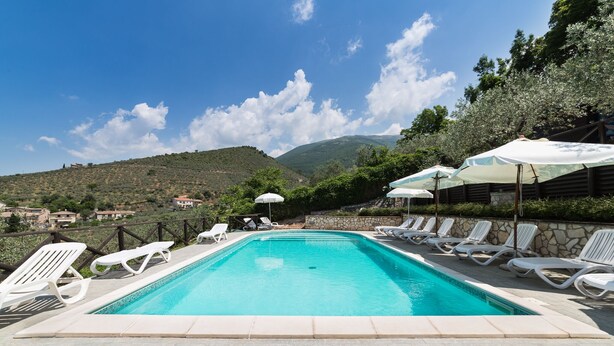 Piscina com cadeiras de descanso brancas e guarda-sóis, cercada por muros de pedra e vista para colinas.
