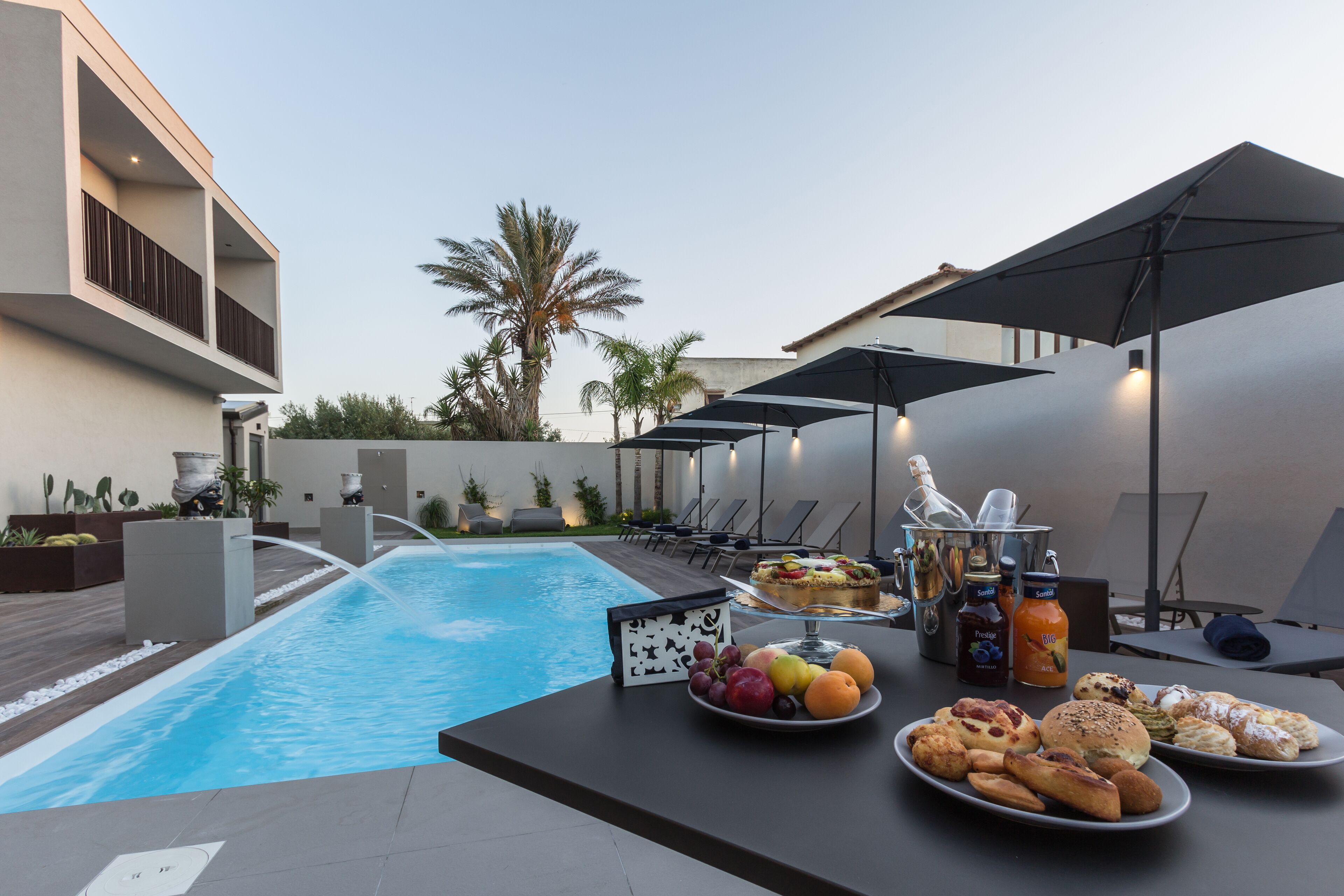 Zona de piscina con mesas dispuestas para comer, mostrando platos con pasteles, frutas, un pastel y bebidas.