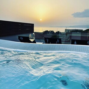 Une piscine sur le toit, avec un verre de vin et un verre d’eau, offrant une vue sur un coucher de soleil se reflétant sur une étendue d’eau.