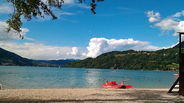 A lake with a red boat on the shore, surrounded by hills and clear skies. - Ap. Annivè – Relaxation and comfort just steps from the heart of Levico Terme (Levico Terme)