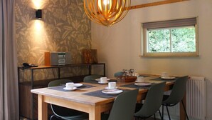 Un coin repas avec une table en bois dressée pour quatre personnes, entourée de chaises. On y trouve une fenêtre donnant sur un paysage verdoyant, un téléphone fixé au mur et un luminaire décoratif au-dessus de la table.