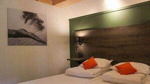 Une chambre d’hôtel avec une tête de lit en bois, deux lits avec une literie blanche et des oreillers orange, une table de chevet et une lampe fixée au mur.