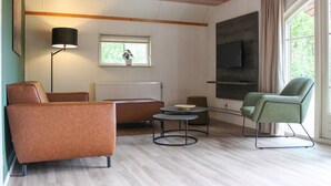 Un salon moderne avec un canapé, un fauteuil et une petite table ronde. On y trouve une fenêtre donnant sur un espace verdoyant, un radiateur et un téléviseur fixé au mur.