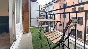 Een balkon met een houten tafel en stoelen, uitzicht op omliggende gebouwen, en een deur die naar binnen leidt.
