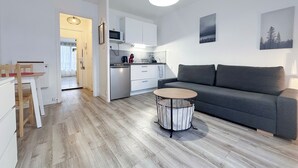 Een modern appartement met een grijze bank, een ronde salontafel, een keuken met witte kasten, en een eetgedeelte met een houten tafel en stoelen.