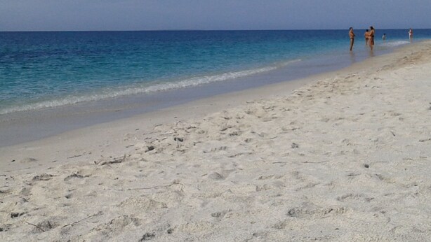 Ein Sandstrand mit klarem, blauem Wasser und ein paar Personen, die am Ufer entlanggehen.