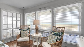 A room with wicker chairs, a small round table with a lamp, and large windows with blinds. - Spacious Oceanfront home near golf and pool, BHI (Bald Head Island)