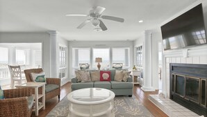 A spacious living room with a ceiling fan, a fireplace, and a large window with blinds. - Spacious Oceanfront home near golf and pool, BHI (Bald Head Island)