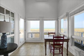 A room with a table and chairs, large windows with a view of the beach, and a lamp on a table.