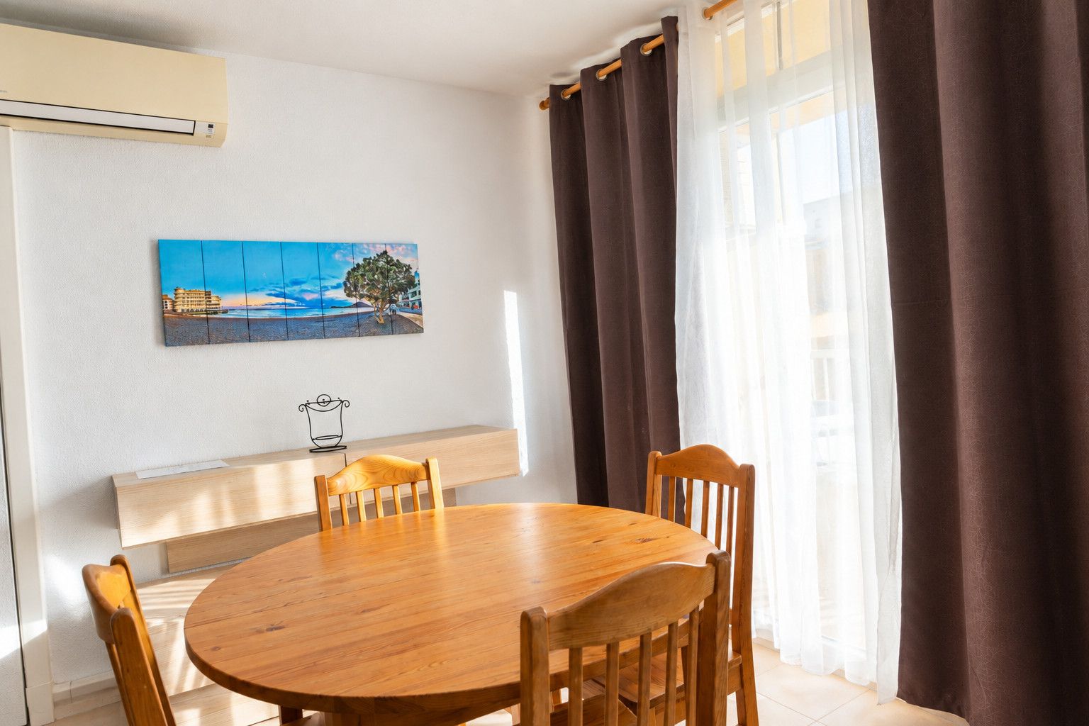 Un comedor con una mesa redonda de madera, cuatro sillas, un aire acondicionado instalado en la pared, un cuadro con una escena costera y una ventana con cortinas marrones.