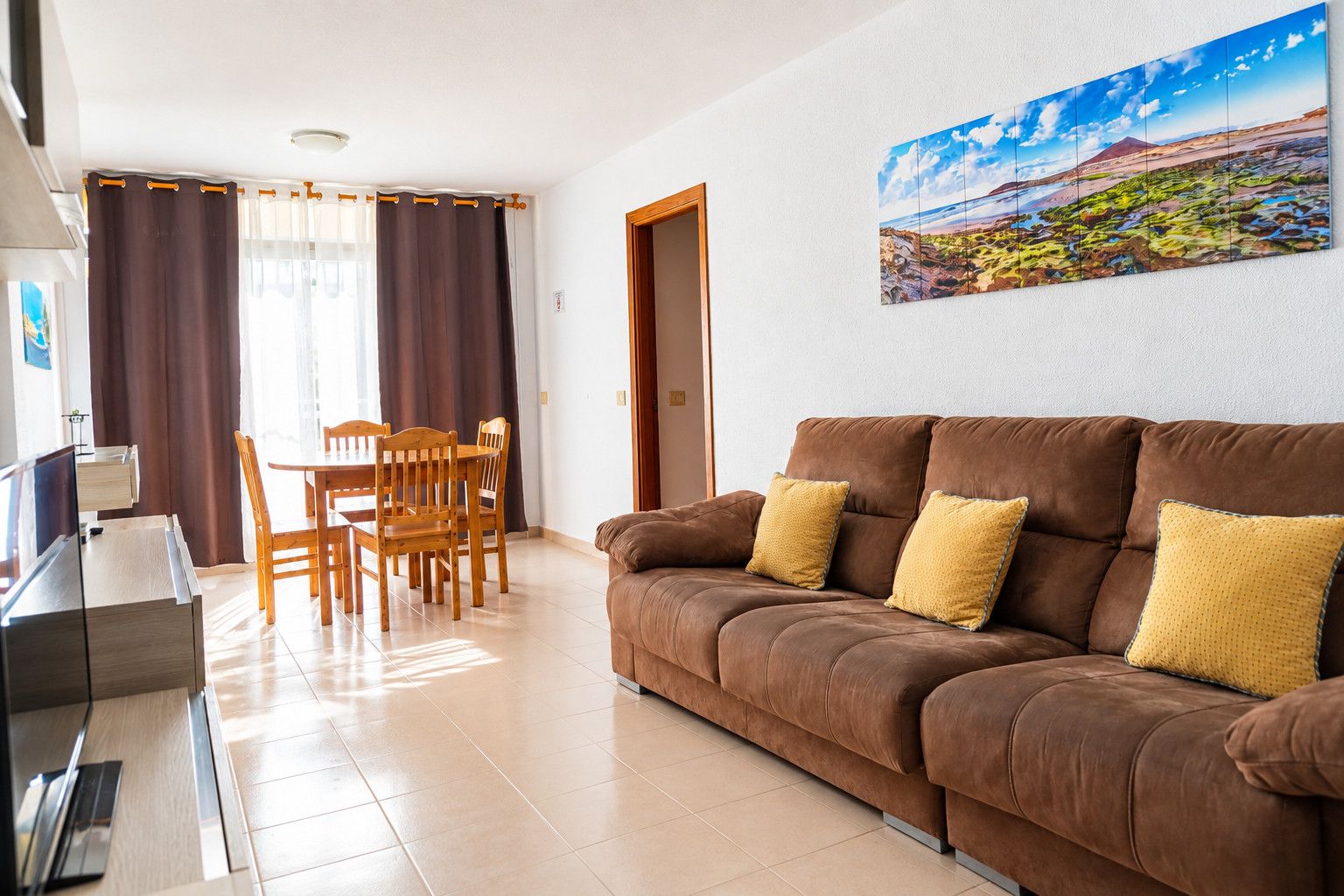 Una sala de estar con un sofá marrón, cojines amarillos, una mesa de comedor con sillas y un cuadro en la pared.