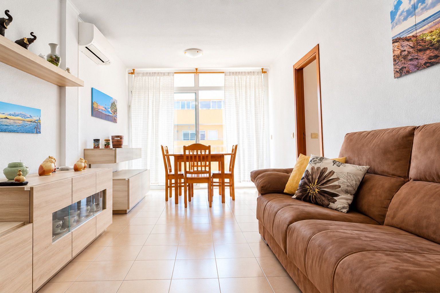 Una sala moderna con un sofá de cuero marrón, una mesa de comedor con sillas, una zona de cocina con armarios y un ventanal con cortinas.
