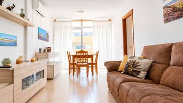 Una sala moderna con un sofá de cuero marrón, una mesa de comedor con sillas, una zona de cocina con armarios y un ventanal con cortinas.