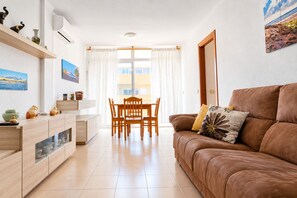 En moderne stue med en brun lædersofa, et spisebord med stole, et køkkenområde med skabe og et stort vindue med gardiner.