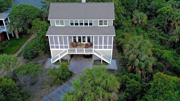A two-story house with a covered porch and a deck, surrounded by dense greenery.