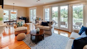 A spacious living room with a wooden floor, a ceiling fan, a dining area with a table and chairs, and a balcony with a view of trees.