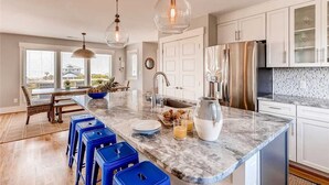 A modern kitchen with a marble countertop, stainless steel appliances, and blue bar stools. - Oceanview. Steps to South Beach • Stunning Sunset Views • Golf Carts Included • (Bald Head Island)