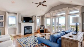 A spacious living room with a fireplace, a large sofa, and a view of the outdoors. - Oceanview. Steps to South Beach • Stunning Sunset Views • Golf Carts Included • (Bald Head Island)