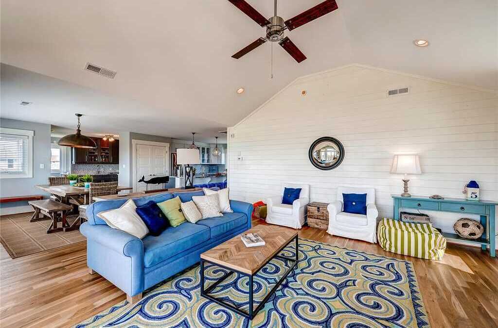 A spacious living area with a blue sofa, white armchairs, a coffee table, and a patterned rug. A ceiling fan and a round mirror are visible on the wall.