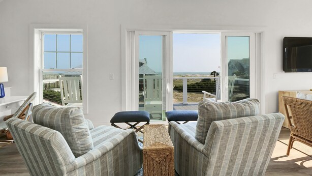 A living room with two striped armchairs, a small table, and a wicker ottoman. There are large windows and a TV mounted on the wall.