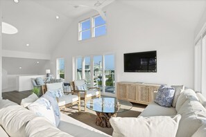 A spacious living room with a high ceiling, large windows, a round glass coffee table, and a comfortable seating area with sofas and chairs.