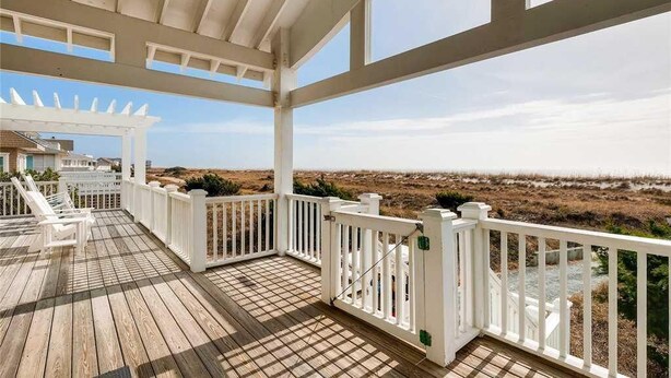 Una terraza de madera con barandillas blancas y una pérgola, con vista a una playa.