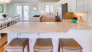 Una cocina con armarios blancos, una isla central con taburetes, un refrigerador y una ventana.