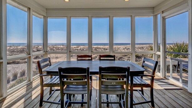 Ein überdachter Essbereich im Freien mit Tisch und Stühlen, große Fenster mit Blick auf den Strand.