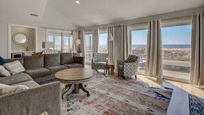 Ein geräumiges Wohnzimmer mit einer großen Couch, einem runden Couchtisch und gemusterten Teppichen. Mehrere Fenster mit Vorhängen bieten einen Blick ins Freie.