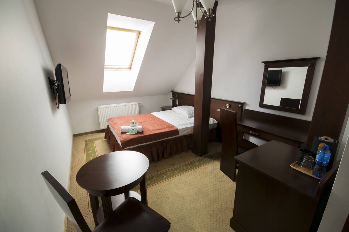 Habitación de hotel compacta con cama, una mesita, una silla, escritorio, espejo y radiador.