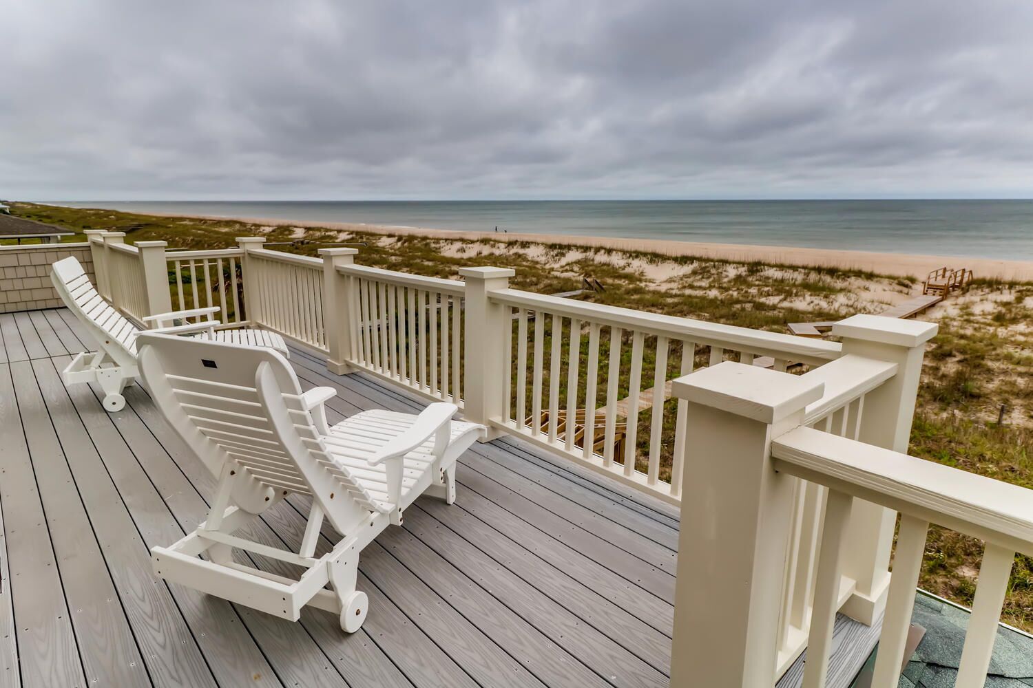 Eine Holzterrasse mit weißen Stühlen und einem Geländer mit Blick auf einen Strand.
