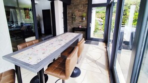 A modern dining area with a long table, leather chairs, a stone wall, and a view of greenery outside. - Beautiful apartment for up to 6 people, south-facing terrace, beach on foot! (La Tremblade)