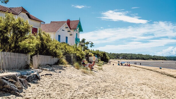 A beach with a white building with red and green accents, a wooden fence, and people relaxing on the sand. - Beautiful apartment for up to 6 people, south-facing terrace, beach on foot! (La Tremblade)
