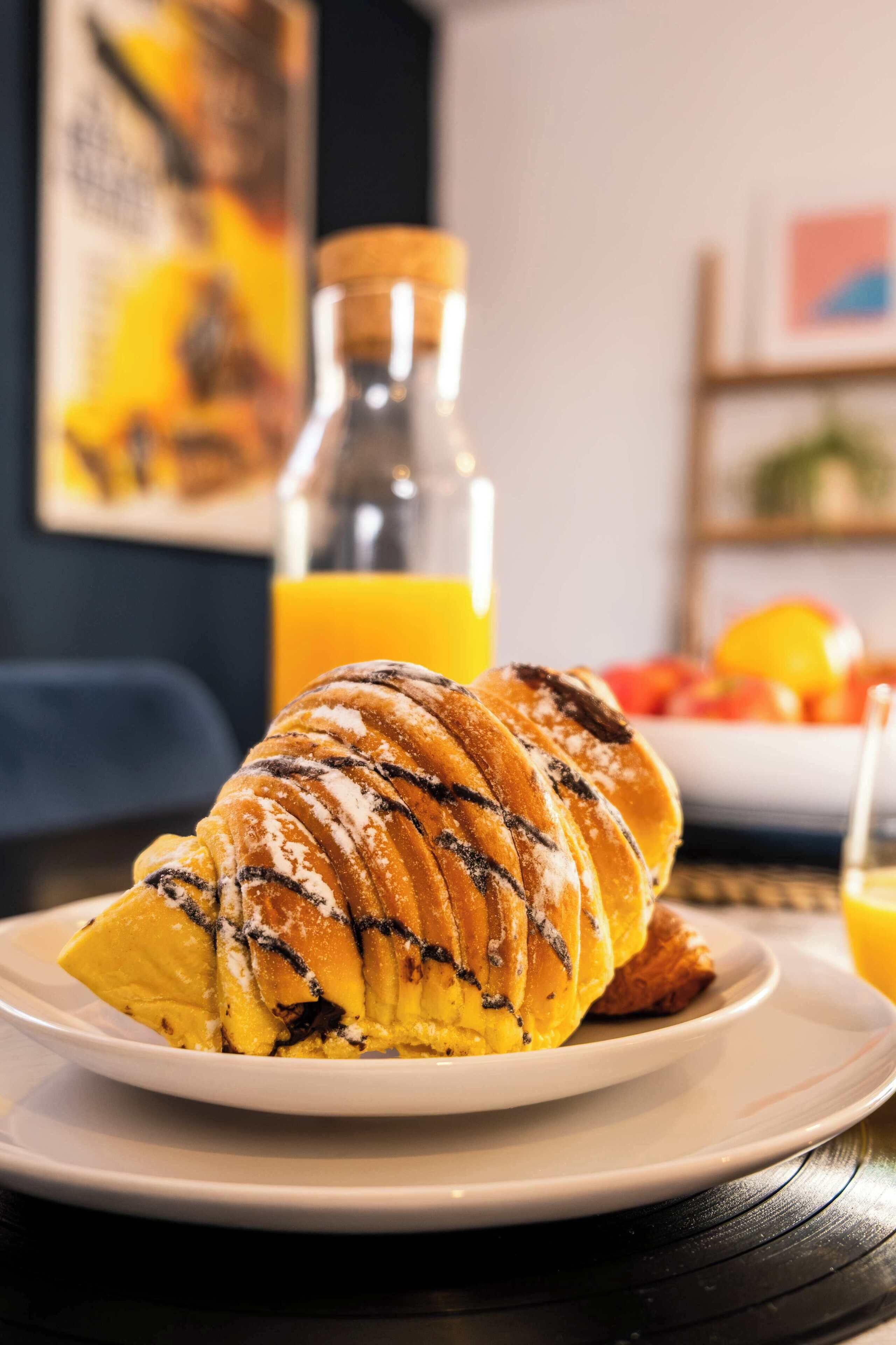 Een croissant op een bord, met een fles sinaasappelsap op de achtergrond.