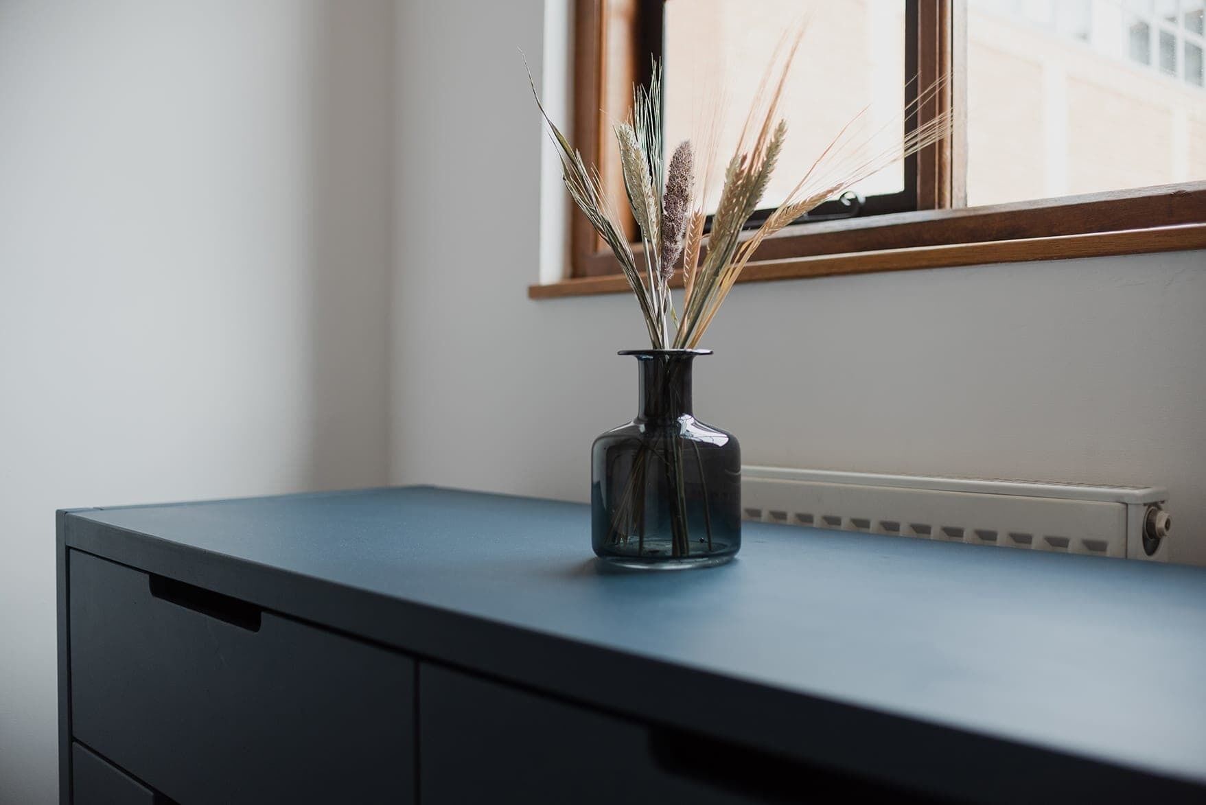 A dark blue desk with a glass vase containing dried plants.
