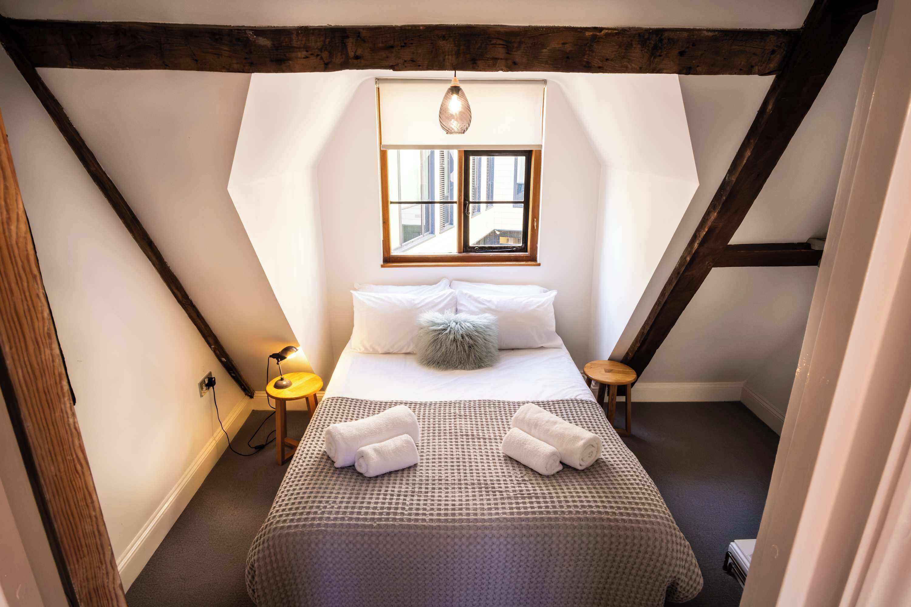 Een slaapkamer met een schuine kap, een bed met een geborduurde spreideken, twee nachtkastjes met lampen en een raam met jaloezieën.