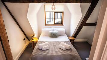 Een slaapkamer met een schuine kap, een bed met een geborduurde spreideken, twee nachtkastjes met lampen en een raam met jaloezieën.