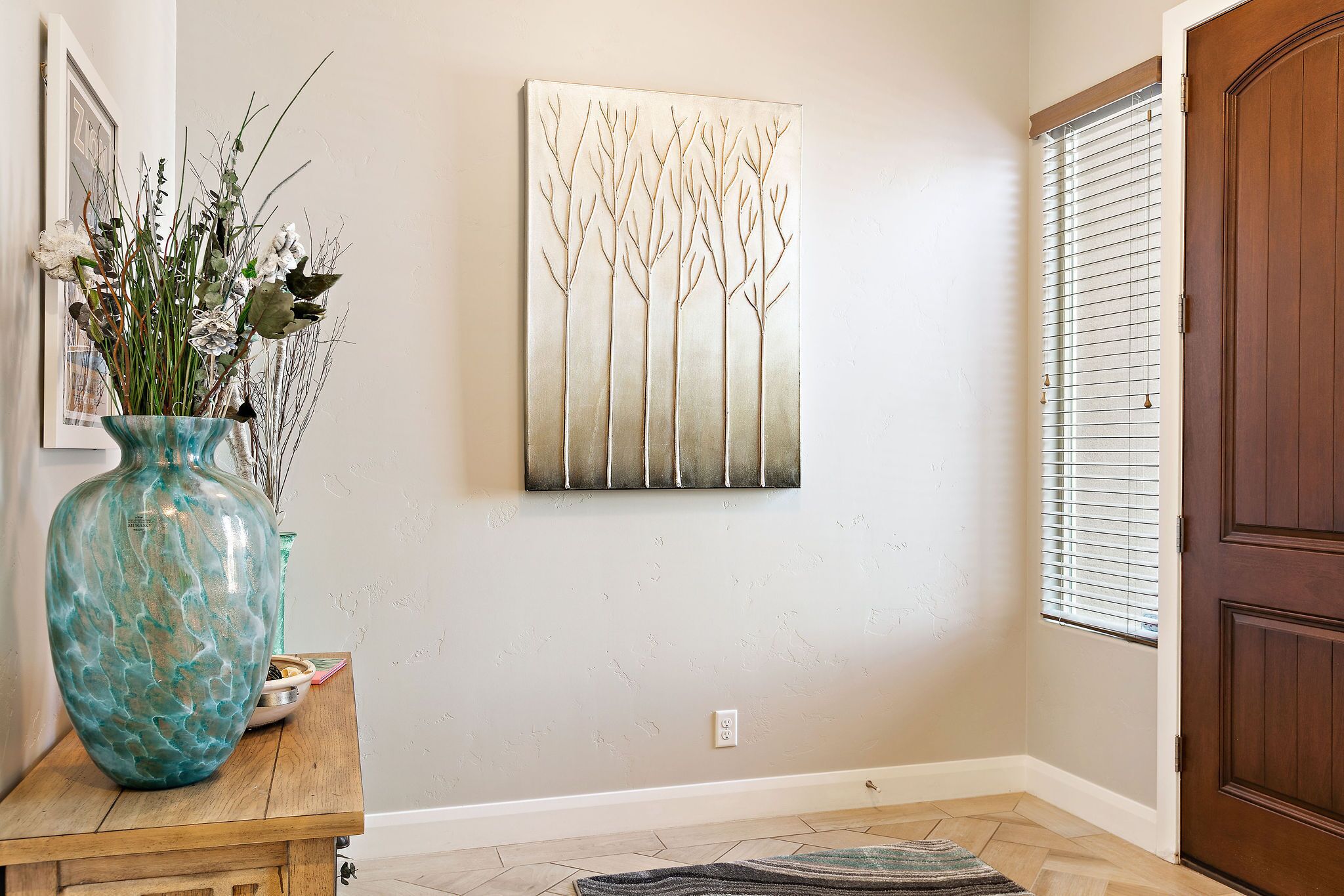 A hallway with a wooden floor, a large vase with dried plants, a framed picture, and a door with blinds.