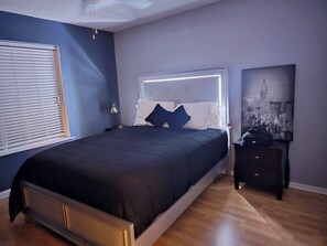 Ein Schlafzimmer mit einem Bett, einem Nachttisch, einer Lampe, einem Fenster mit Jalousien und einem Stadtbild an der Wand.